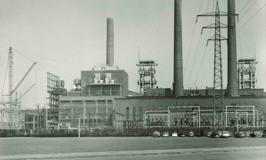 Black and white photo of an industrial power plant with three tall smokestacks, electrical towers, and several parked cars in front. Two signs on the building read WATTSUN and POWER.
