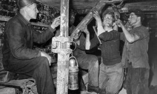 A group of coal miners, faces and clothes covered in soot, work and inspect machinery in an underground mine. One miner sits to the left, while others kneel and use tools under low ceilings, illuminated by a hanging lantern.