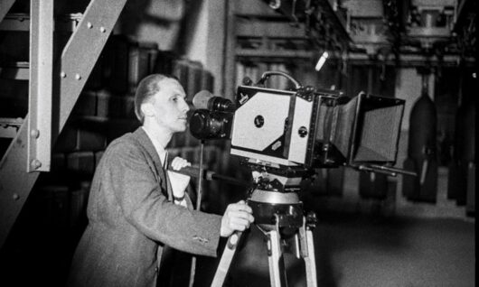 Een man in pak bedient binnenshuis een ouderwetse filmcamera die op een statief is gemonteerd. Op de achtergrond hangen industriële apparatuur en grote objecten.