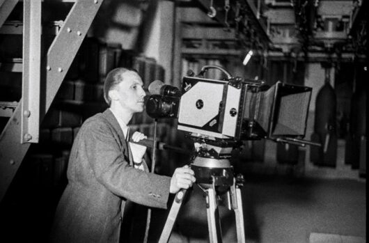 A man in a suit operates a vintage film camera mounted on a tripod indoors, with industrial equipment and large objects hanging in the background.