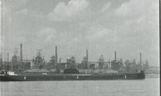 Zwart-witfoto van een industriële staalfabriek met hoge schoorstenen en machines aan de waterkant; op de voorgrond ligt een grote binnenvaartboot aangemeerd. Rechtsonder staat 'Historisch Archief Krupp'.