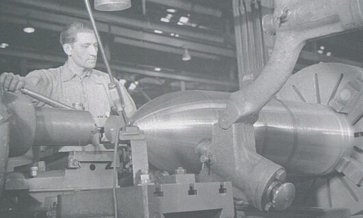 Een man bedient zware machines en vormt een groot metalen object in een industriële fabriek. De werkruimte staat vol met apparatuur en de verlichting is gericht op het werkgebied.