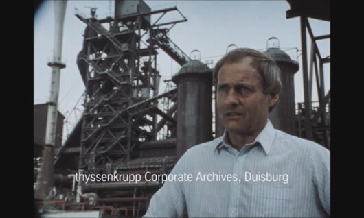 A man in a light blue shirt stands in front of an industrial steel plant with large towers and metal structures. The text reads thyssenkrupp Corporate Archives, Duisburg.