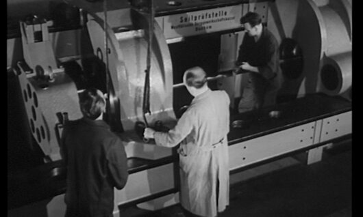 Three men work around a large industrial machine in a factory setting; one wears a lab coat while the others are in work clothes, all focused on operating or inspecting the equipment.