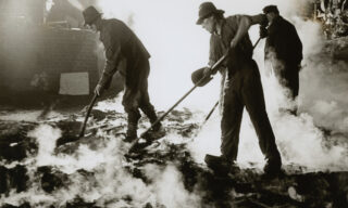 Three workers in protective clothing and helmets use shovels to move smoking slag inside a steel mill. Bright light and steam surround them, highlighting the intense industrial environment.
