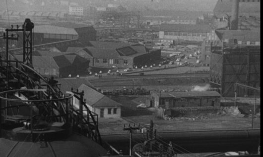 Een zwart-witfoto toont een industrieel complex met diverse grote fabrieksgebouwen, opslagtanks en spoorrails. De achtergrond is wazig en er staan ​​talloze voertuigen tussen de gebouwen geparkeerd.