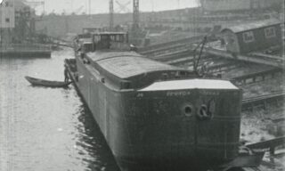 Black and white image of a large cargo ship docked at an industrial shipyard, surrounded by cranes and buildings, with a smaller boat alongside. The scene appears historical and the weather is overcast.