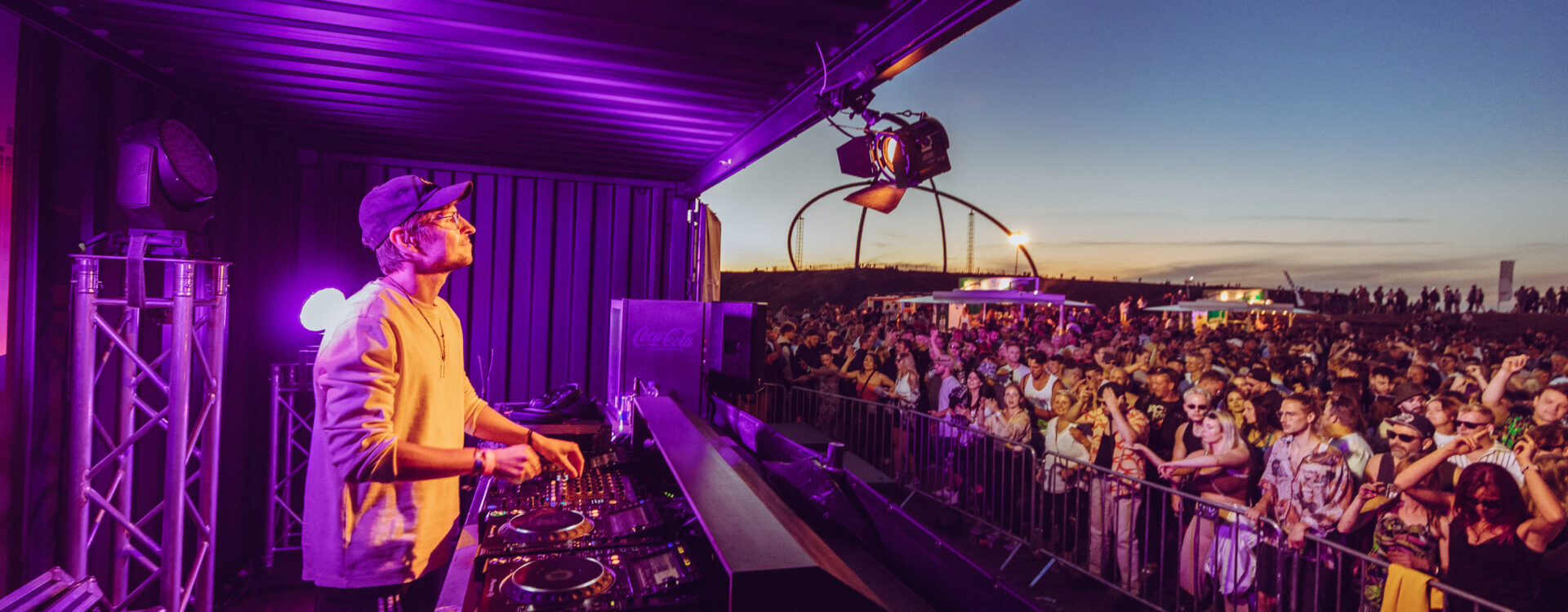 A DJ wearing headphones performs on stage at an outdoor music festival at sunset as a large crowd dances and enjoys the event. Bright lights and festival setups are visible in the background.