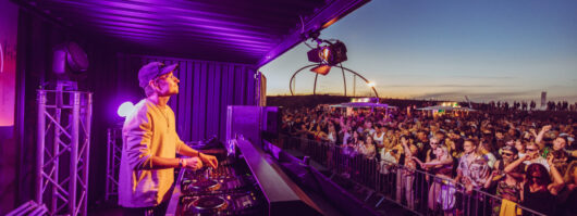 A DJ wearing headphones performs on stage at an outdoor music festival at sunset as a large crowd dances and enjoys the event. Bright lights and festival setups are visible in the background.