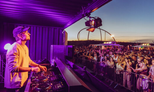A DJ wearing headphones performs on stage at an outdoor music festival at sunset as a large crowd dances and enjoys the event. Bright lights and festival setups are visible in the background.