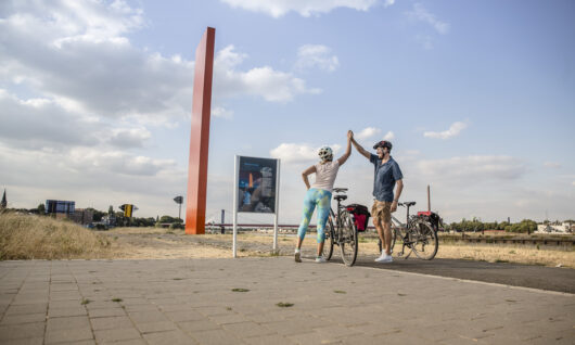 Two cyclists stand next to their bikes on a paved path, giving a high five near a tall red sculpture and a sign, with a partly cloudy sky and grassy landscape in the background.