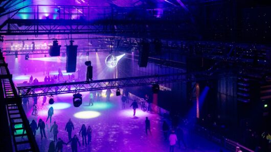 People are ice skating indoors under vibrant purple and blue lights, with a disco ball hanging from the ceiling and spotlights creating colorful patterns on the rink. The space has a festive, lively atmosphere.