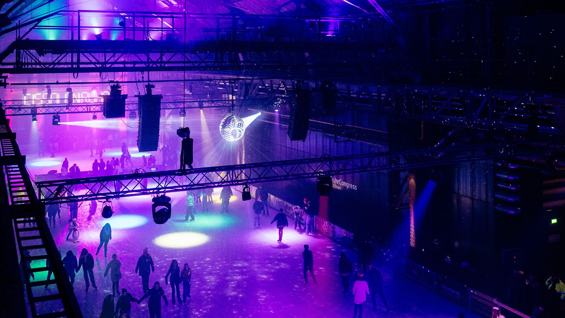 People are ice skating indoors under vibrant purple and blue lights, with a disco ball hanging from the ceiling and spotlights creating colorful patterns on the rink. The space has a festive, lively atmosphere.