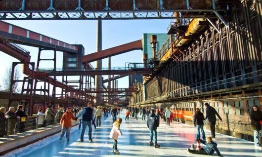 People of all ages ice skating outdoors on a long rink surrounded by industrial structures and pipes, with bright sunlight and blue sky in the background. Some skaters are moving while others stand or sit on the ice.