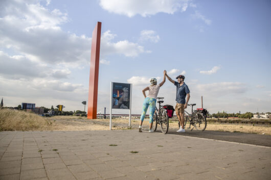 Two cyclists stand beside their bikes on a paved path, giving a high five near a tall red sculpture and a sign, with a partly cloudy sky and grassy landscape in the background.